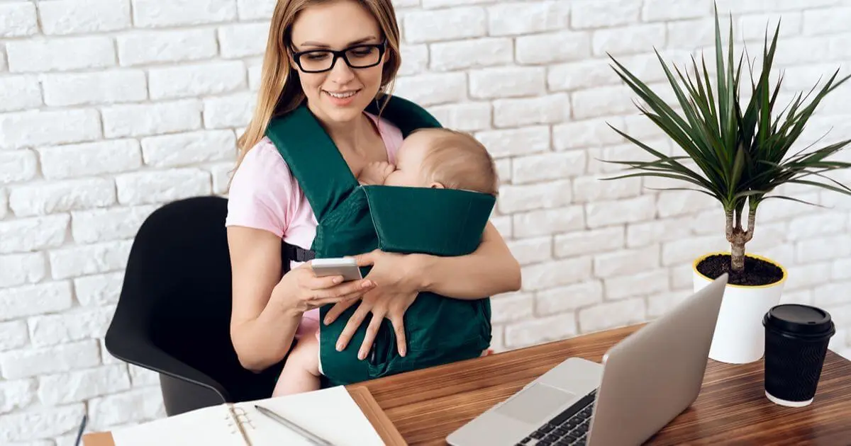 Returning to Work: Mom wearing a baby carrier while checking her phone at her home office before returning to work.