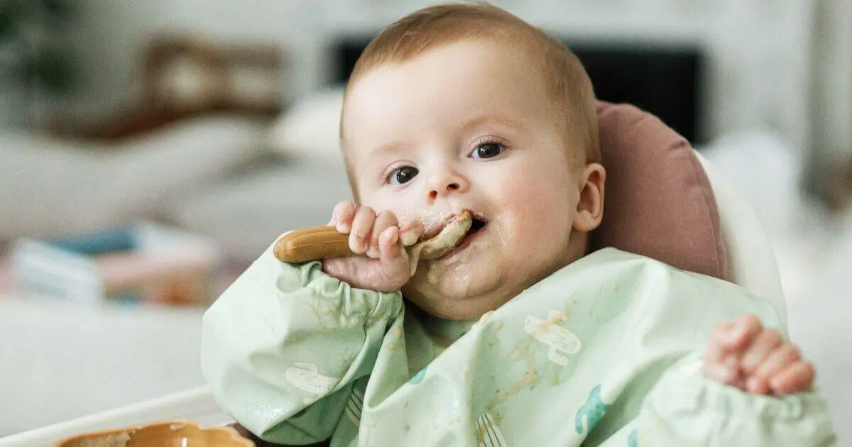 Introducing Solids Step by Step: What, When, and How to Feed Your Baby Adorable messy baby in high chair during introducing solids, trying porridge for the first time after six months.
