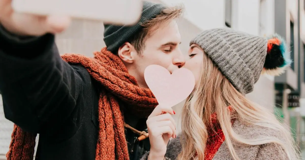 Couple sharing a loving moment while taking a heart-shaped selfie, celebrating Valentine’s Day with a baby in a warm, intimate setting.