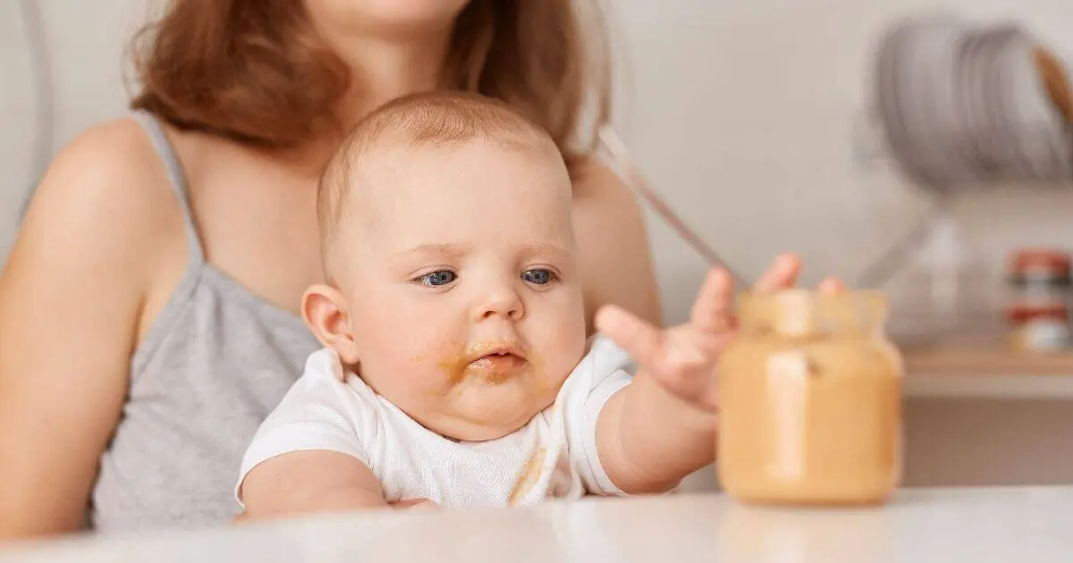 Introducing Solids Step by Step: What, When, and How to Feed Your Baby Curious baby reaching toward food jar while mother feeds vegetable purée during introducing solids at home.