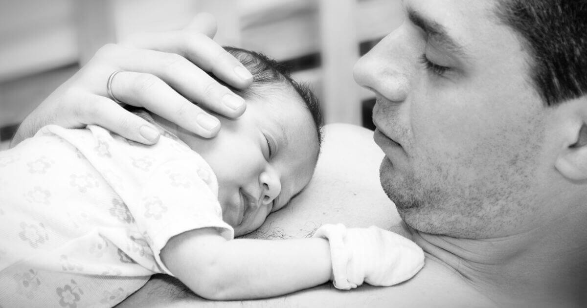 A cute blue-eyed newborn baby lying calmly while a father gently holds the baby’s tiny hand, capturing a tender moment of early fatherhood.