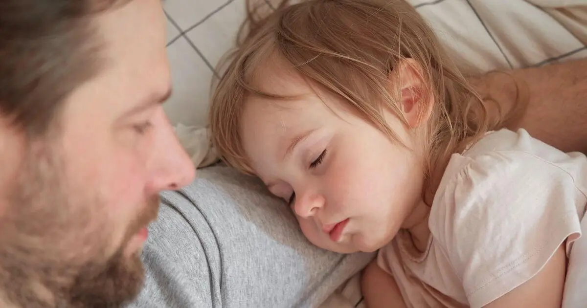 A father sleeping peacefully next to his toddler daughter in a white bed at home, capturing a quiet and loving fatherhood moment in everyday family life.