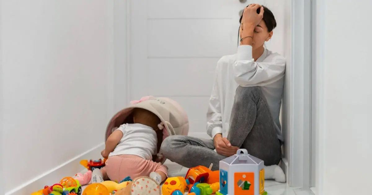 Mother resting against the wall as her baby plays nearby, capturing a quiet moment often associated with mom guilt and mental overload.