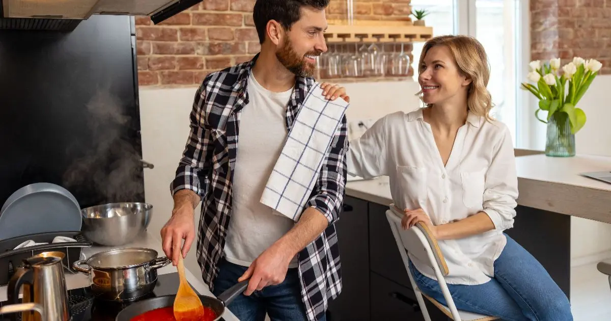 Mental load shared between partners as a father cooks dinner while a mother relaxes, showing healthy responsibility sharing in family life.