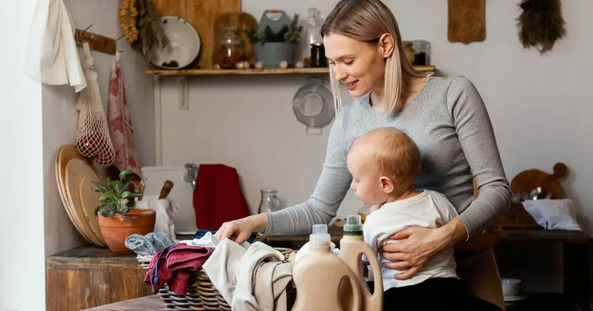 Mother folding laundry while holding her baby in a cozy kitchen, showing everyday motherhood moments often linked to mom guilt.
