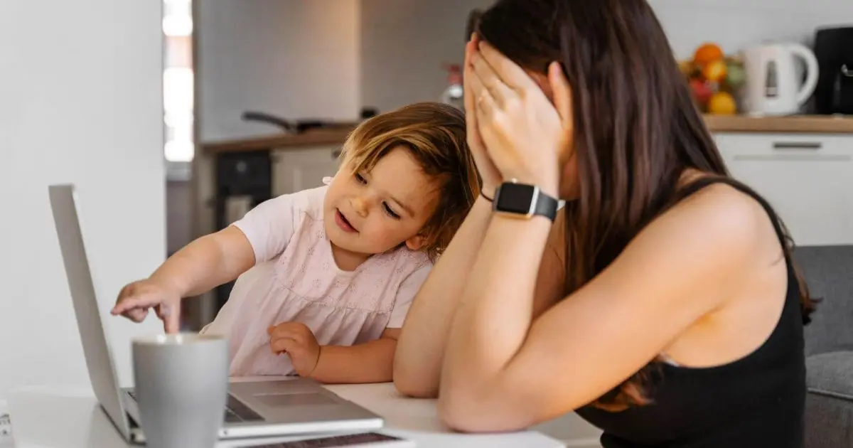 Stressed mother working from home while caring for a crying baby and toddler, showing the emotional and mental load of mom burnout and remote work motherhood.