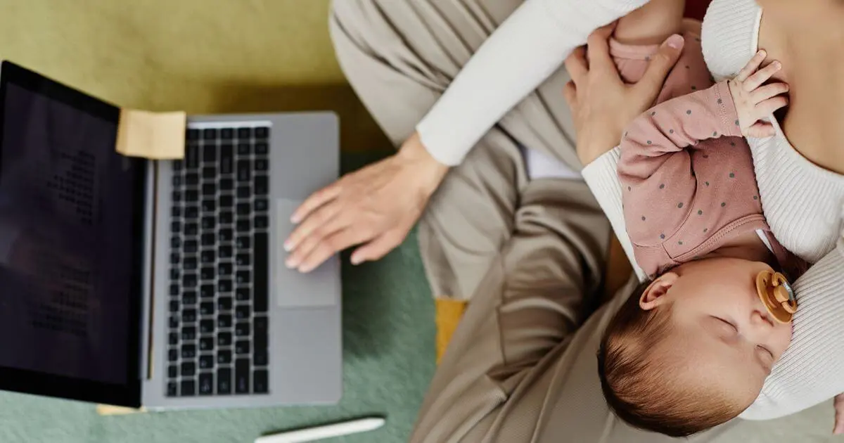 Returning to Work: Overhead view of a mother working on her laptop while holding her sleeping baby after returning to work.