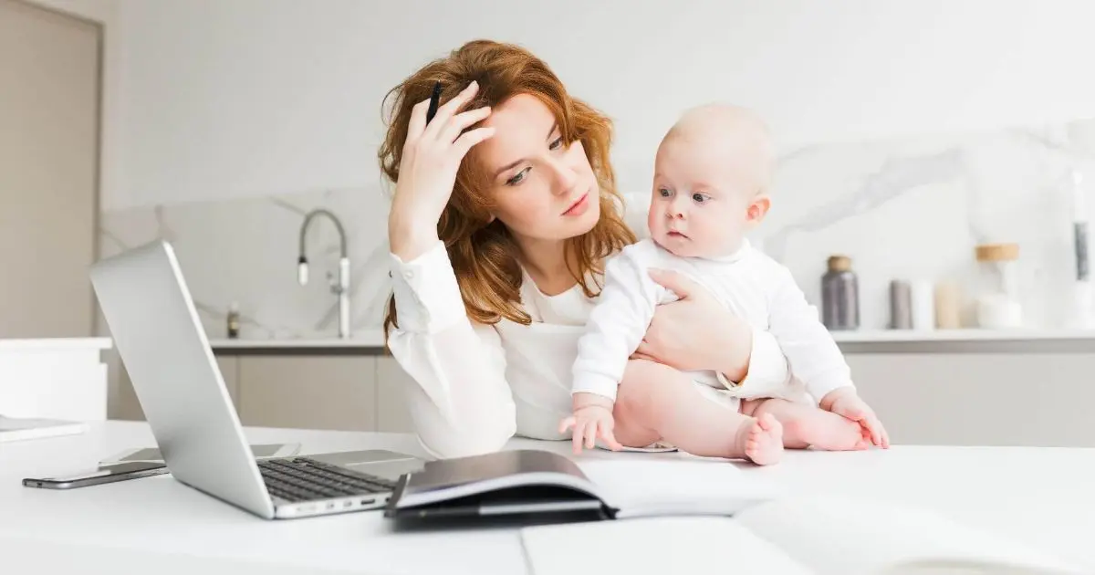 Mom burnout awareness — thoughtful working mother holding her baby while sitting at a table, balancing motherhood, work, and emotional responsibility.
