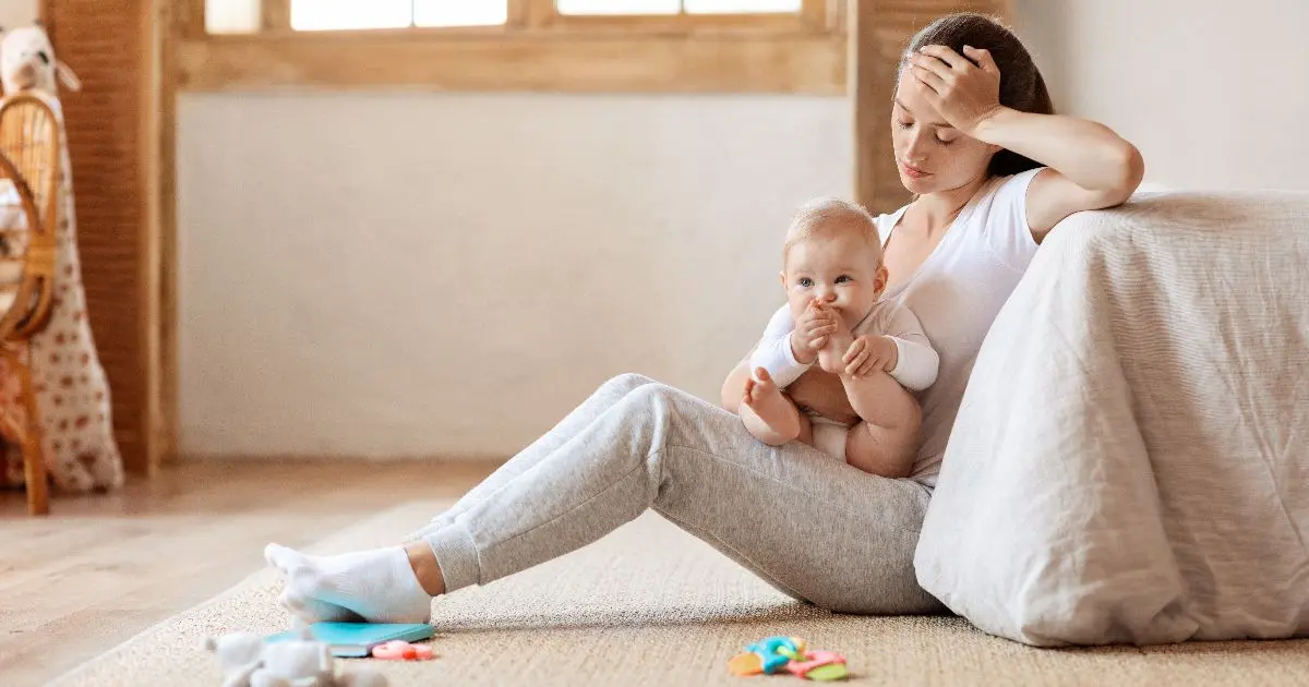 Mom Burnout in Summer: Tired mother sitting on the floor with her baby, feeling overwhelmed by parenting responsibilities, mental load, and the exhaustion that often comes with summer motherhood.