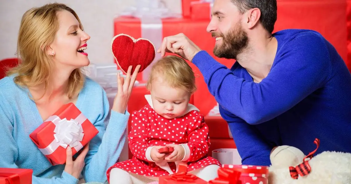 Happy family with mother, father, and young daughter holding gift boxes at home, celebrating Valentine’s Day with a baby in a loving, joyful atmosphere.