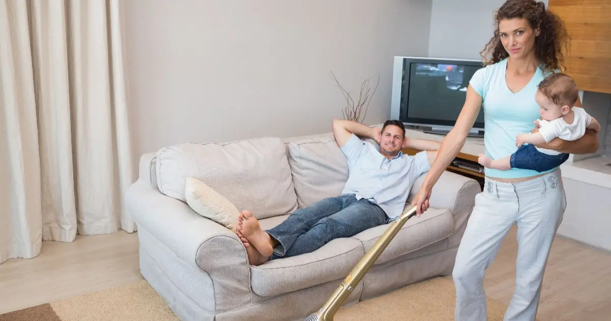Mental load in everyday motherhood as a mom vacuums while holding her baby, while her partner relaxes on the couch, illustrating the invisible workload and emotional burden many mothers carry.