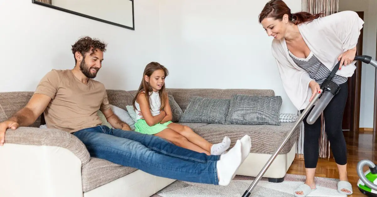 Mental load in family life as a mom does the housework while her partner and daughter relax, illustrating the invisible workload many mothers carry every day.