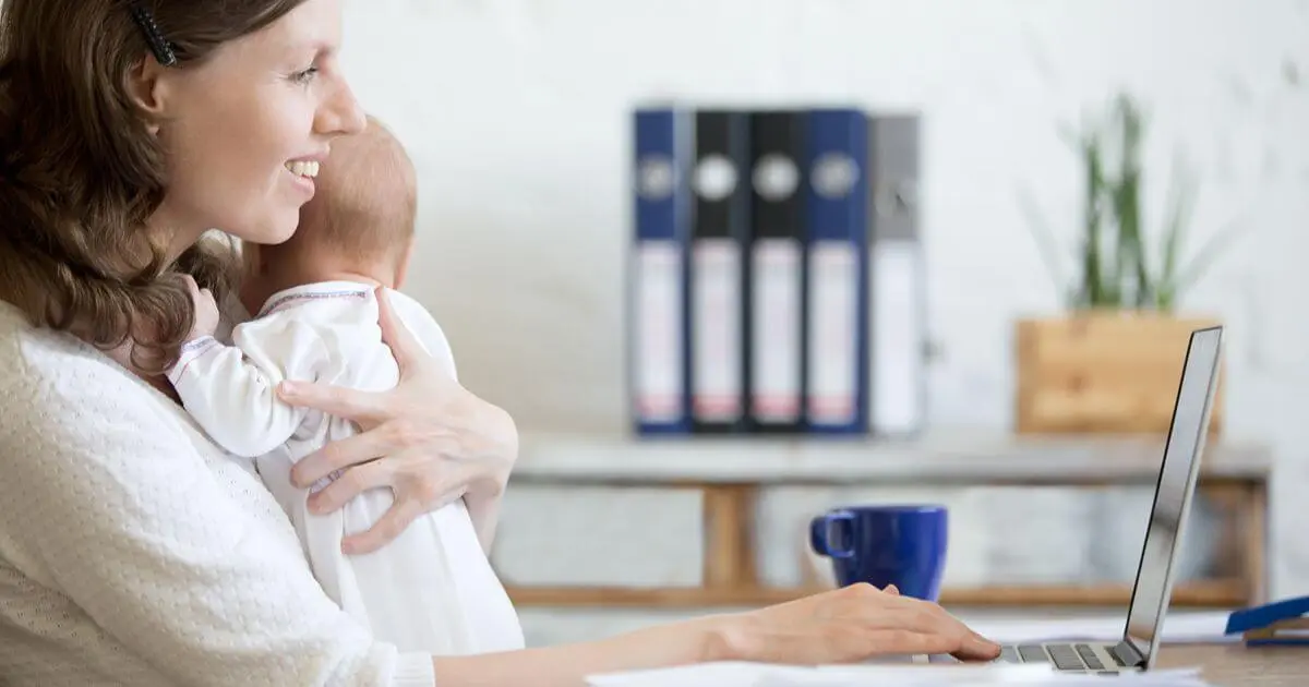 Returning to Work: Smiling mother holding her baby while working on a laptop from home during her transition back to work.
